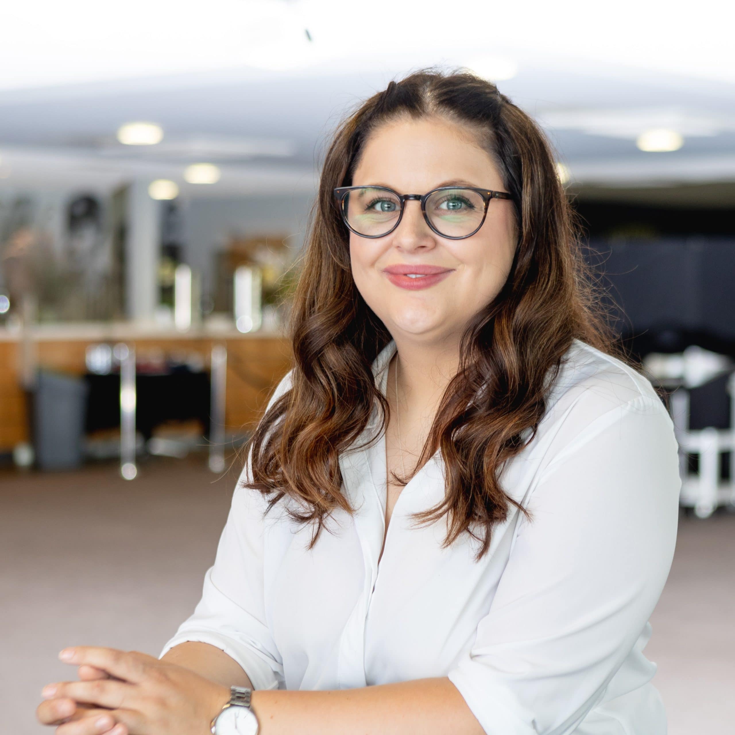 A brunette feminine person with curly long hair, wearing a white shirt and black glasses.